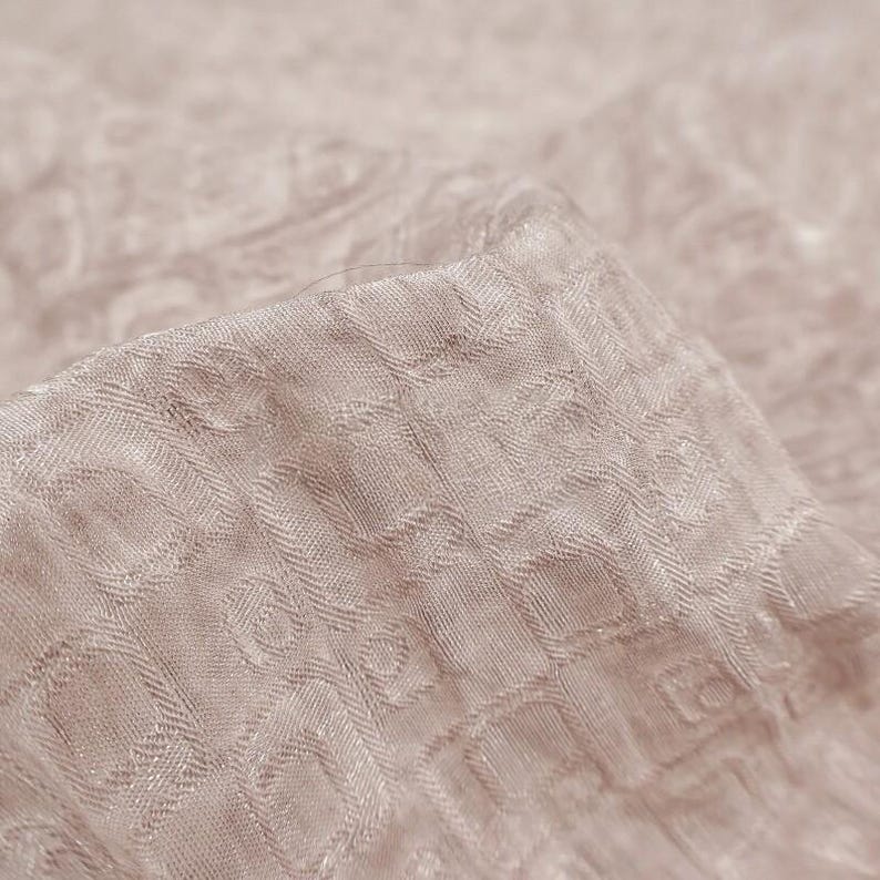 May include: Close-up of a folded piece of fabric in a light beige color. The fabric has a textured pattern of small, repeating geometric shapes. The material appears soft and slightly shiny, with subtle variations in tone.