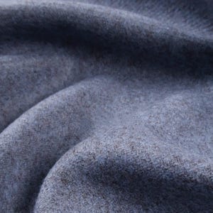 May include: Close-up of a textured, blue-grey fabric with subtle colour variations. The material appears soft, possibly woollen, with visible fibres and a slightly mottled appearance. The fabric is draped, creating shadows and highlights.