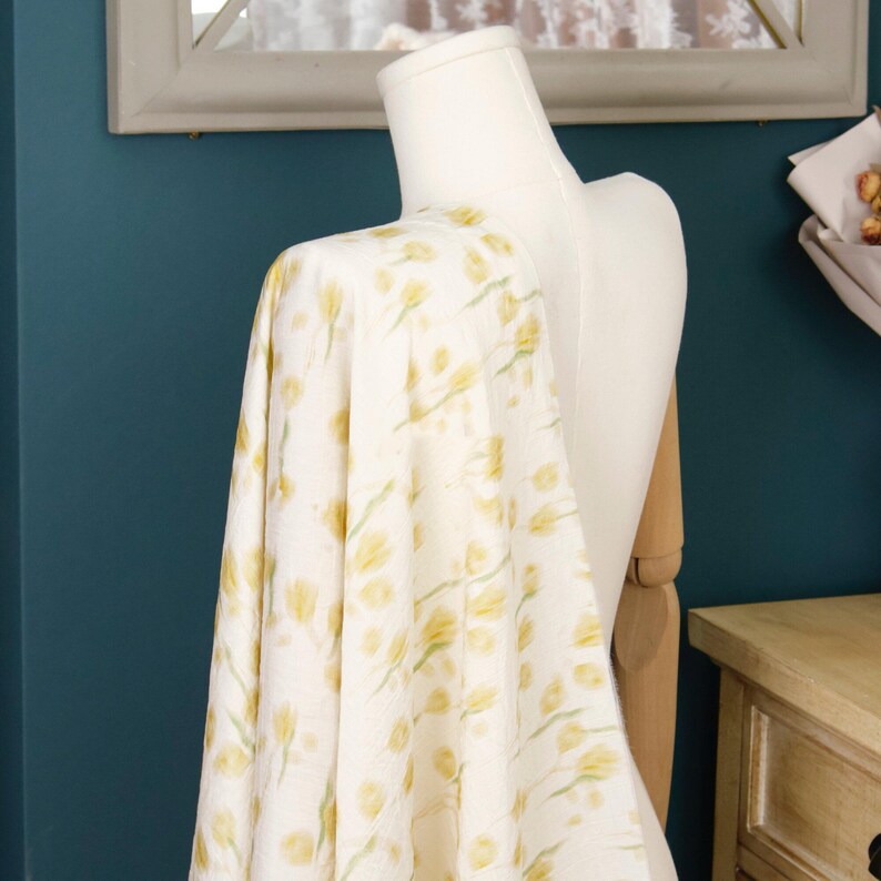 May include: A piece of off-white fabric draped over a mannequin, featuring a repeating pattern of soft yellow tulips with light green stems and leaves. The textile has a textured, slightly crinkled appearance throughout the entire surface.