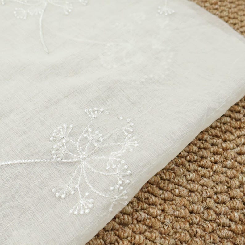 May include: Close-up of a white fabric with delicate, embroidered dandelion-like floral patterns. The fabric has a slightly sheer quality, with the embroidered details adding texture and visual interest. The background is a textured, brown surface.