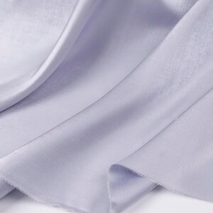 May include: Close-up of a flowing, light purple fabric with a subtle sheen. The fabric is draped in soft folds, creating a sense of movement and texture. The edges of the fabric are slightly frayed, adding a touch of rustic charm.