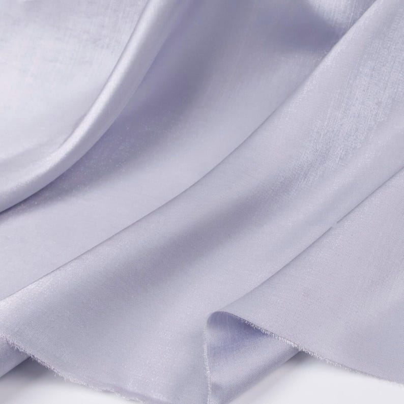 May include: Close-up of a flowing, light purple fabric with a subtle sheen. The fabric is draped in soft folds, creating a sense of movement and texture. The edges of the fabric are slightly frayed, adding a touch of rustic charm.
