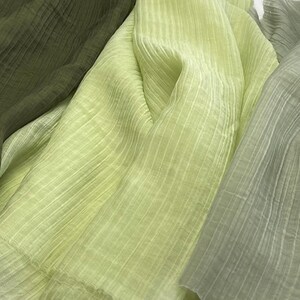 May include: Close-up of layered fabric in shades of green. The fabric has a crinkled texture with vertical lines. The colors range from a deep olive green to a pale, almost white-green, creating a soft, textured appearance.