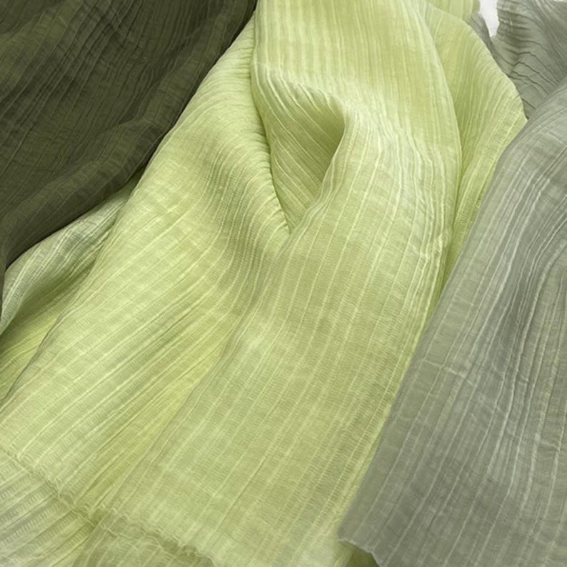 May include: Close-up of layered fabric in shades of green. The fabric has a crinkled texture with vertical lines. The colors range from a deep olive green to a pale, almost white-green, creating a soft, textured appearance.
