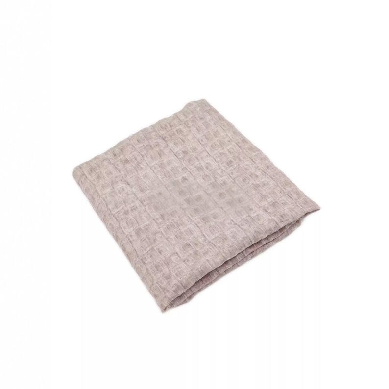 May include: A folded, light beige waffle-weave blanket. The blanket is square and appears to be made of a soft, textured material. The weave pattern creates a grid-like design across the surface. The blanket is on a white background.