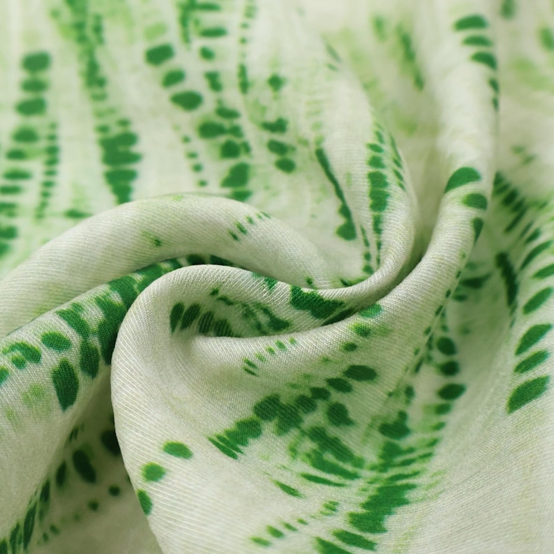 Op de afbeelding: Een lichtgewicht stof met een tie-dye patroon met levendige groene vlekken en strepen in verticale lijnen op een zachte gebroken witte achtergrond. Het materiaal is in plooien gelegd, wat de textuur en de herhalende print benadrukt.