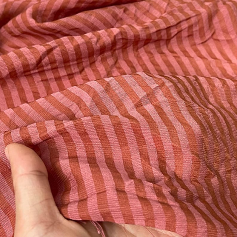 Peut inclure: Un tissu l&eacute;ger pr&eacute;sentant un motif r&eacute;p&eacute;titif de fines rayures altern&eacute;es dans des tons orange terre cuite et rose tendre. Le mat&eacute;riau poss&egrave;de une texture froiss&eacute;e visible avec une qualit&eacute; semi-transparente, cr&eacute;ant un effet visuel superpos&eacute;.