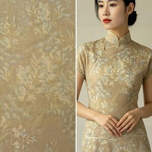 Puede incluir: Un vestido de manga corta en tono bronceado con un sutil estampado floral en tonos azul apagado y crema. La prenda tiene un cuello alto tipo mandar&iacute;n con un detalle de cierre diagonal en el pecho, acabado con ribetes delicados en las costuras.