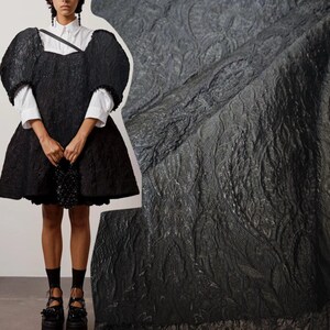 May include: A black textured dress with a square neckline and voluminous puff sleeves worn over a white collared shirt. The dress features a raised floral pattern and is paired with black socks and black platform shoes with beaded accents.