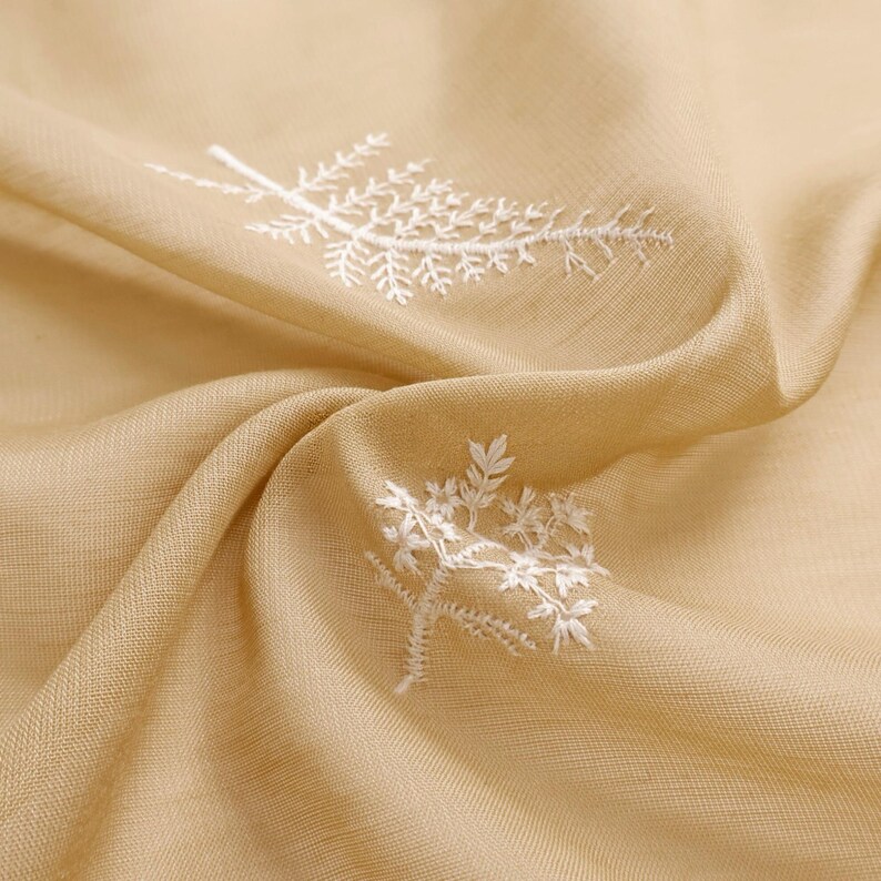 May include: Close-up of a beige fabric with white embroidered floral designs. The fabric is draped, showing folds and texture. The embroidery features delicate, detailed botanical patterns, adding a touch of elegance to the textile.