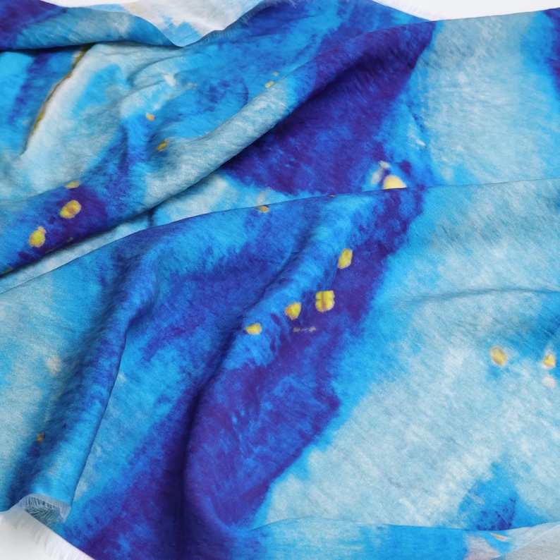 May include: A close-up of a blue and white fabric with a watercolor effect. The fabric has a tie-dye appearance with shades of blue and white, and small yellow dots scattered throughout. The fabric is folded and draped.