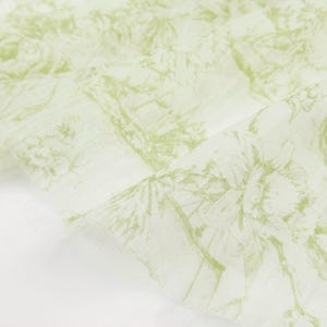 May include: A light green and white fabric with a botanical design. The fabric features a repeating pattern of trees, flowers, and other foliage. The edges of the fabric are frayed.