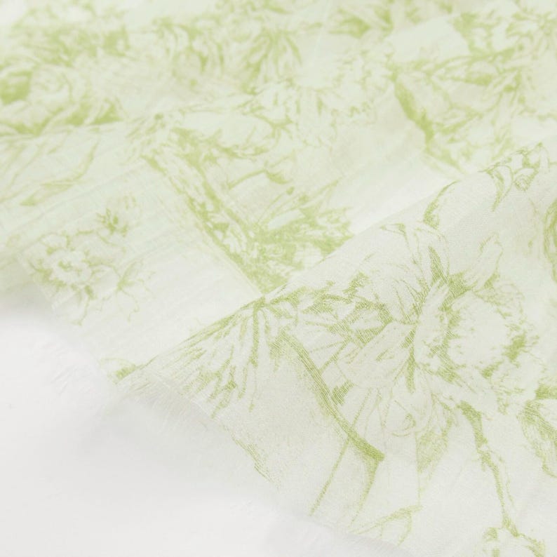May include: A light green and white fabric with a botanical design. The fabric features a repeating pattern of trees, flowers, and other foliage. The edges of the fabric are frayed.