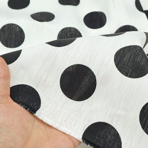 May include: White fabric with large black polka dots. The fabric is held up, showing the texture and pattern. The polka dots are evenly spaced and vary slightly in size. The fabric has a soft, flowing appearance, suitable for clothing or home decor.