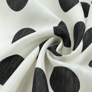 May include: Close-up of a white fabric with a pattern of large, black polka dots. The fabric is slightly textured and appears to be draped, with folds and shadows adding depth to the design. The polka dots vary in size and are irregularly spaced.