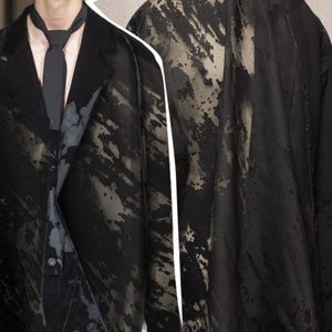Puede incluir: Un blazer negro y una bufanda larga a juego en tela semitransparente con un patrón abstracto, oscuro y moteado. El blazer tiene solapa de muesca y cierre de un solo botón, usado con una corbata negra sólida sobre una camisa de color claro.