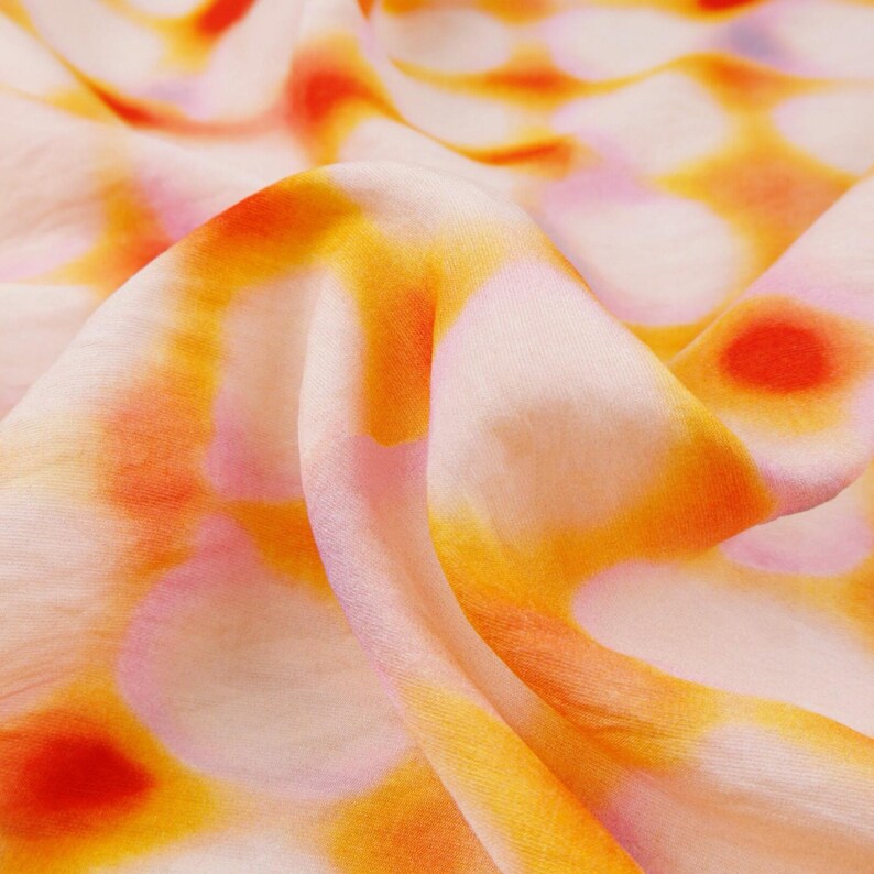 May include: Close-up of a flowing fabric with a pattern of orange, yellow, and red circular shapes on a pale pink background. The fabric is soft and appears to be silk or a similar lightweight material. The design is abstract and artistic.
