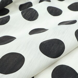 May include: Close-up of white fabric with large, irregularly shaped black polka dots. The fabric is slightly wrinkled, creating shadows and texture. The polka dots vary in size and shape, adding visual interest to the textile.