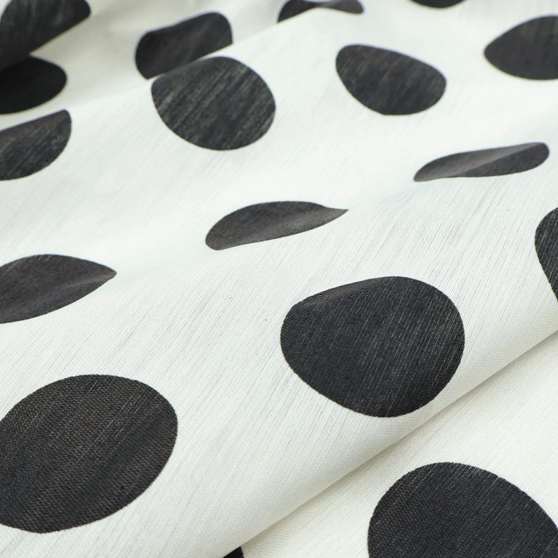 May include: Close-up of white fabric with large, irregularly shaped black polka dots. The fabric is slightly wrinkled, creating shadows and texture. The polka dots vary in size and shape, adding visual interest to the textile.
