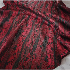 May include: A fabric featuring a deep red and black color scheme with a textured, crinkled surface. The material displays alternating vertical stripes of solid black and intricate, ornate floral patterns in red, creating a layered visual effect.