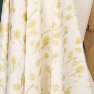 May include: A piece of off-white fabric featuring a repeating pattern of soft yellow tulip-like flower heads and light green stems. The textile has a subtle, crinkled texture throughout, creating a draped effect with visible folds and shadows.