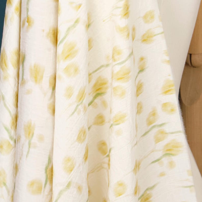 May include: A piece of off-white fabric featuring a repeating pattern of soft yellow tulip-like flower heads and light green stems. The textile has a subtle, crinkled texture throughout, creating a draped effect with visible folds and shadows.
