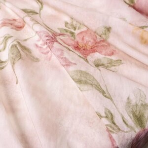 May include: Close-up of a light pink fabric with a floral pattern. The design features watercolor-style flowers in shades of pink and red, with green leaves and stems. The fabric is draped, creating soft folds and shadows.