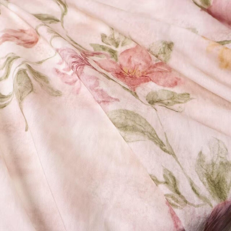 May include: Close-up of a light pink fabric with a floral pattern. The design features watercolor-style flowers in shades of pink and red, with green leaves and stems. The fabric is draped, creating soft folds and shadows.