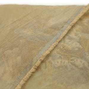 May include: Close-up of a section of fabric with a floral pattern in muted tones of beige, light blue, and hints of orange. The edge of the fabric has a frayed, textured trim, and a thin blue stripe.