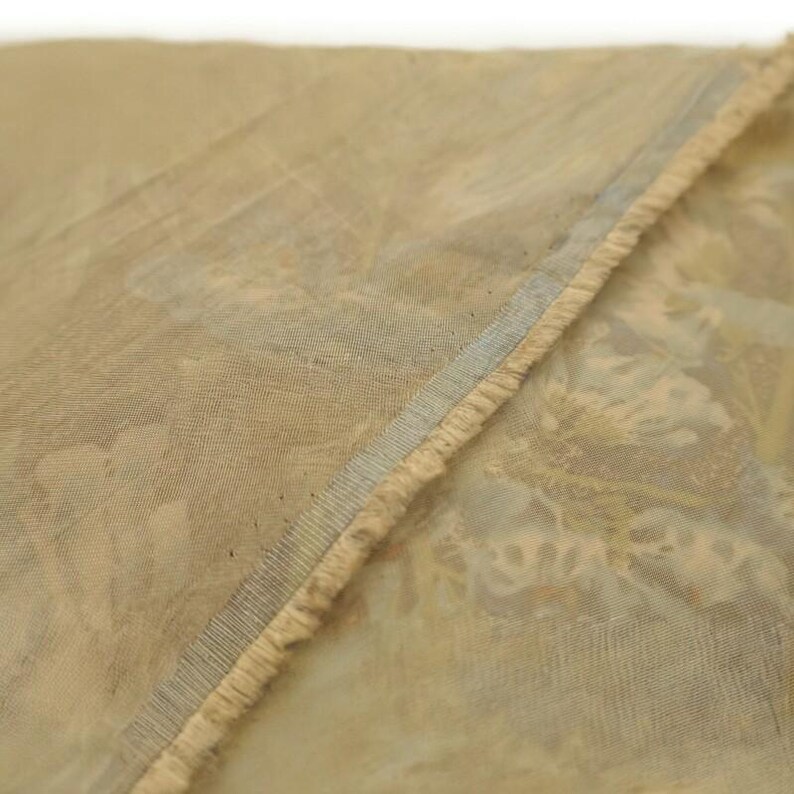 May include: Close-up of a section of fabric with a floral pattern in muted tones of beige, light blue, and hints of orange. The edge of the fabric has a frayed, textured trim, and a thin blue stripe.