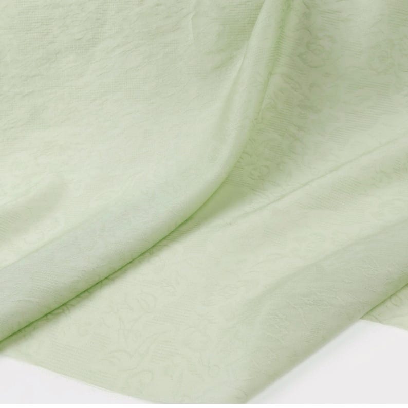 May include: A close-up of a light green fabric with a subtle floral pattern. The fabric is draped, showing folds and textures. The color is a soft, pastel green, and the material appears to be lightweight and flowing.