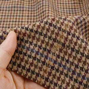 Pode incluir: Um tecido entrelaçado com um padrão clássico de pé-de-galinha numa paleta multicolorida de fios castanhos, bege, vermelho escuro e azul subtil. O têxtil apresenta uma trama visível e texturizada com uma estrutura densa e entrelaçada.