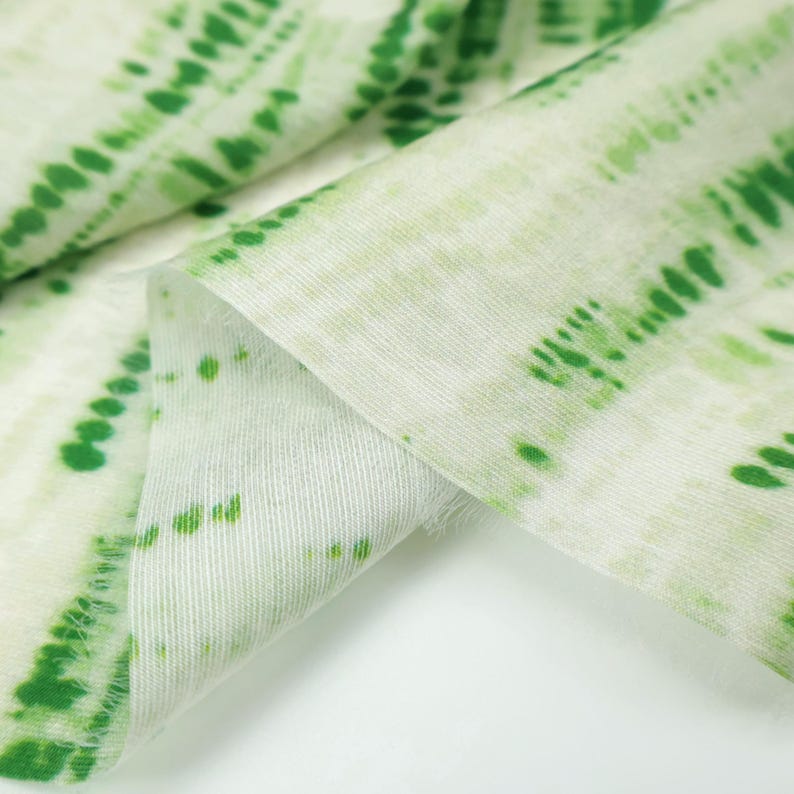 Op de afbeelding: Een stuk stof met een witte basiskleur en een herhalend patroon van verticale, gestippelde groene lijnen. Het textiel heeft een zichtbare geweven textuur en is gevouwen om de diepte van het materiaal en het groene printontwerp te tonen.