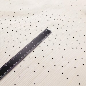 May include: A black ruler measuring up to 19 inches is placed diagonally across a cream-colored fabric. The fabric is patterned with small, evenly spaced black dots. The fabric has a subtle, textured appearance.