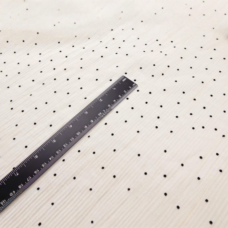 May include: A black ruler measuring up to 19 inches is placed diagonally across a cream-colored fabric. The fabric is patterned with small, evenly spaced black dots. The fabric has a subtle, textured appearance.