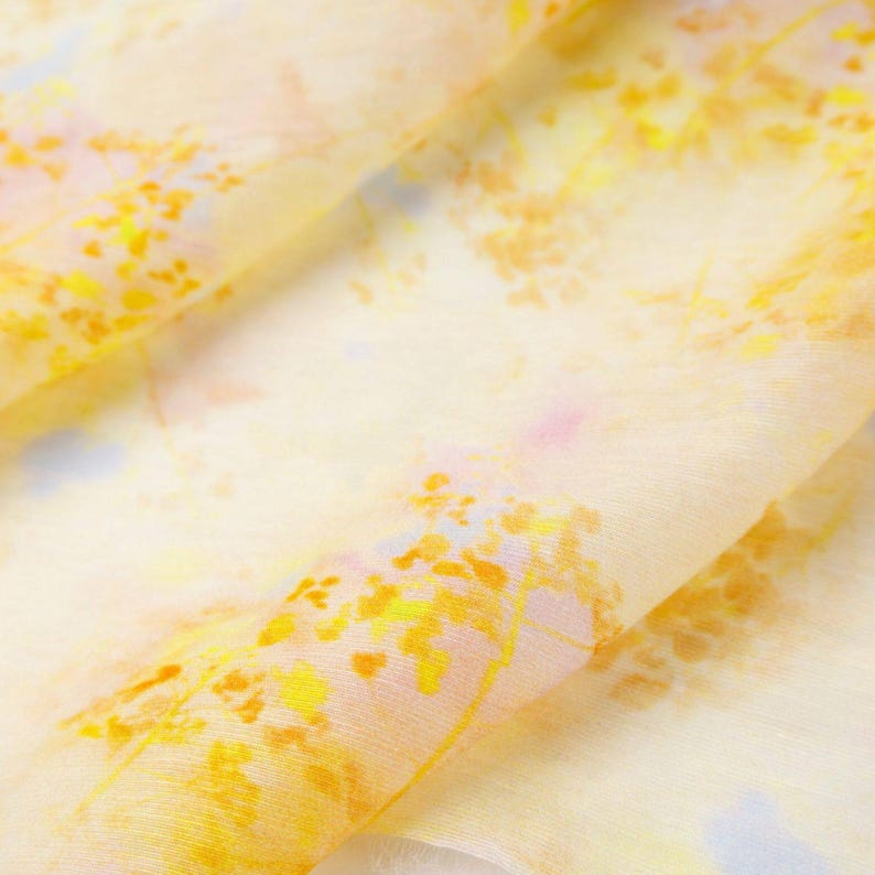 May include: A close-up of a sheer fabric with a watercolor-like design. The fabric features a soft yellow base with accents of yellow, orange, pink, and blue. The design resembles a floral or botanical pattern.
