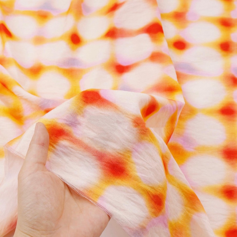 May include: A close-up of a fabric with a repeating pattern of orange, red, and yellow circles on a white background. The fabric is soft and flowing, with visible folds and creases. The pattern is slightly blurred, giving it a watercolor effect.