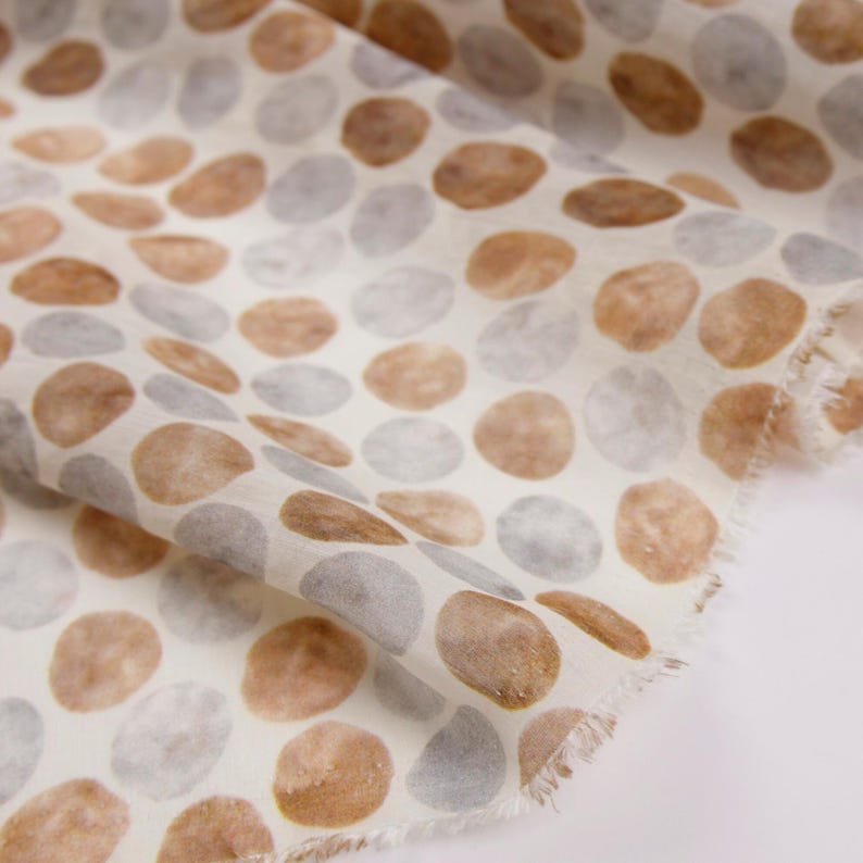 May include: Close-up of a fabric with a repeating pattern of brown and gray oval shapes on a white background. The fabric has a soft, textured appearance, with a frayed edge visible on the right side.