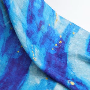 May include: A flowing fabric with a watercolor-like design. The scarf features shades of blue and indigo with accents of yellow. The fabric appears to be lightweight and suitable for a scarf or shawl.