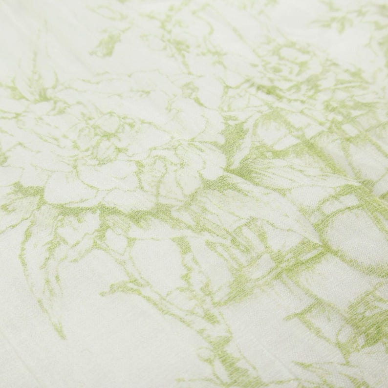 May include: Close-up of a white fabric with a delicate, repeating floral pattern in a soft, muted green. The design appears to be sketched or drawn, creating a textured and artistic effect. The fabric is slightly wrinkled, adding depth.