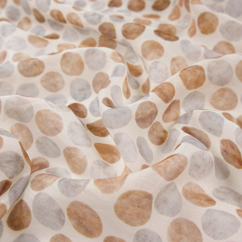 May include: Close-up of a fabric with a pattern of irregular, rounded shapes in shades of brown and gray against a cream background. The fabric is soft and flowing, with visible folds and creases, suggesting a lightweight material.