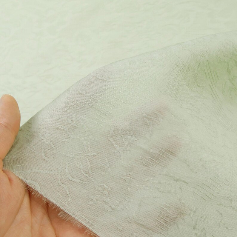 May include: A sheer, pale green fabric with a delicate floral pattern. The fabric is held up, revealing its lightweight and slightly textured nature. The design features subtle, repeating botanical motifs, creating a soft, airy aesthetic.