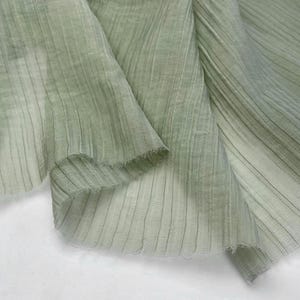May include: Close-up of a light green fabric with vertical ribbed texture. The fabric is draped, showing folds and a frayed edge. The material appears lightweight and semi-transparent, with subtle variations in color.