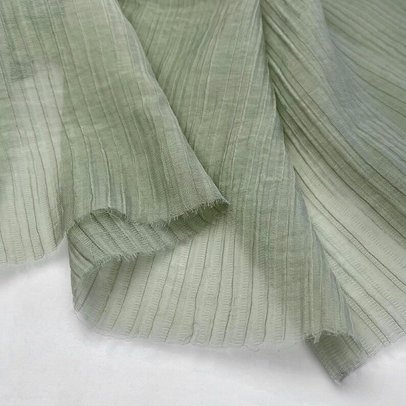 May include: Close-up of a light green fabric with vertical ribbed texture. The fabric is draped, showing folds and a frayed edge. The material appears lightweight and semi-transparent, with subtle variations in color.