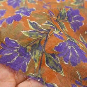May include: Close-up of a floral fabric featuring large purple flowers and green leaves on a burnt orange background. The fabric has a soft, slightly sheer texture, with visible fibers. The design is a repeating pattern of stylized flowers and foliage.