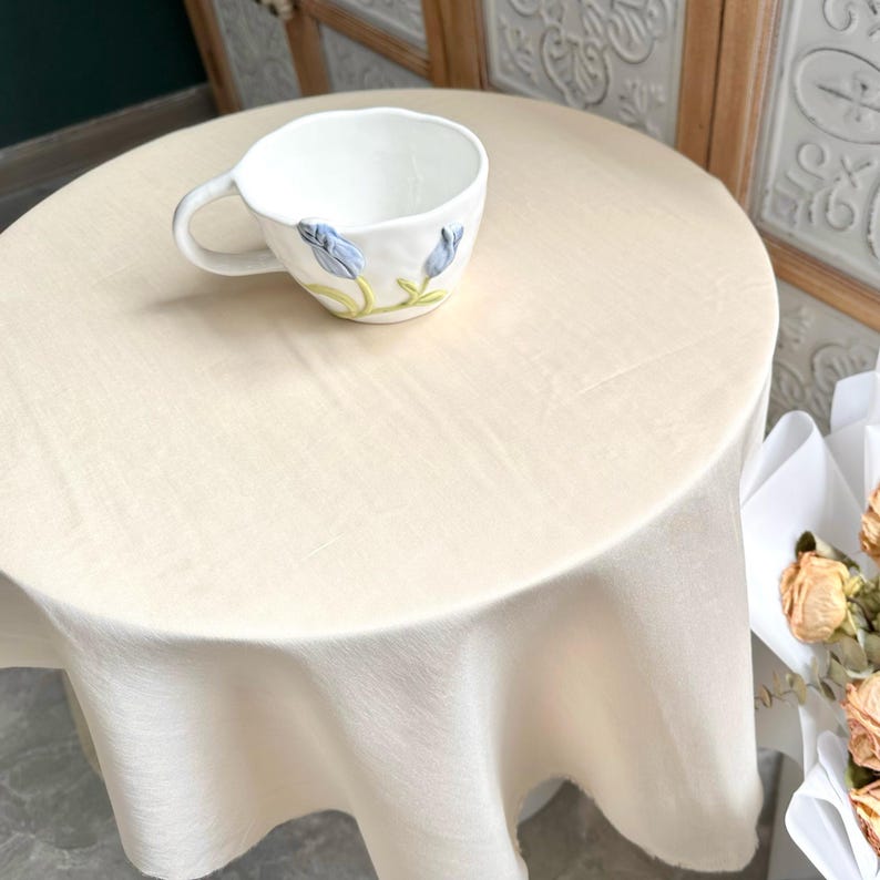 以下が含まれることがあります： 青い花の装飾と緑の茎が施された、白いセラミックマグカップ。 マグカップは、お揃いのテーブルクロスで覆われた丸いクリーム色のテーブルの上に置かれています。 柔らかく自然な光が差し込むシーンです。