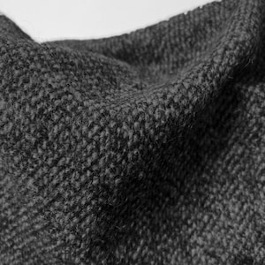 May include: Close-up of a dark gray knitted fabric. The texture is dense and appears to be made of thick yarn, with visible loops and a slightly uneven surface. The fabric is folded, creating shadows and highlighting the texture.