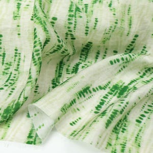 Op de afbeelding: Een lichtgewicht stof met een witte basis en verticale, abstracte groene tie-dye strepen. Het patroon bestaat uit groene stippen en streepjes van verschillende groottes, gerangschikt in vloeiende, onregelmatige kolommen over het semi-transparante textieloppervlak.
