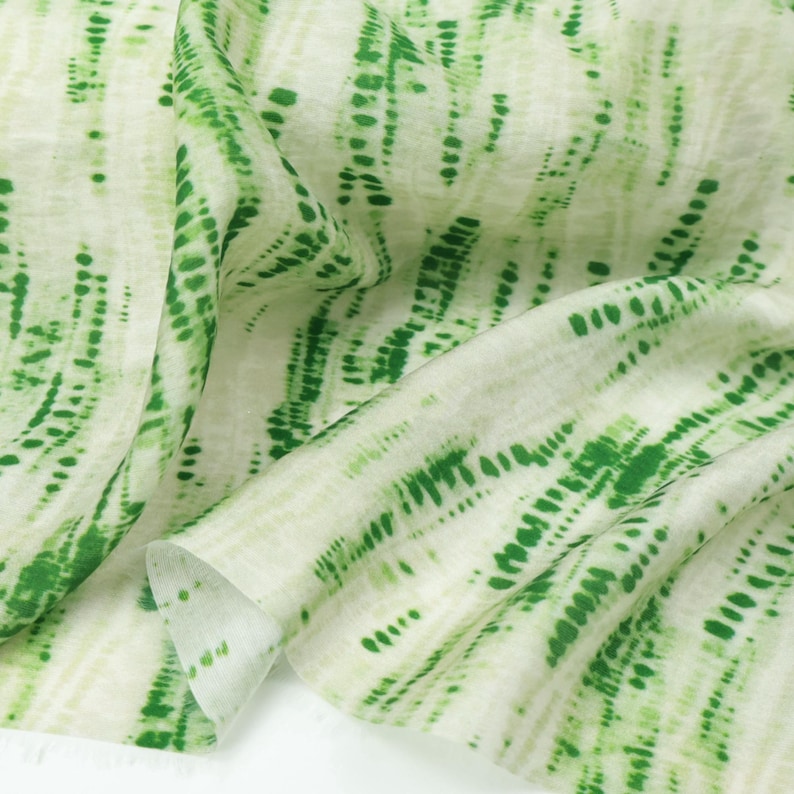 Op de afbeelding: Een lichtgewicht stof met een witte basis en verticale, abstracte groene tie-dye strepen. Het patroon bestaat uit groene stippen en streepjes van verschillende groottes, gerangschikt in vloeiende, onregelmatige kolommen over het semi-transparante textieloppervlak.