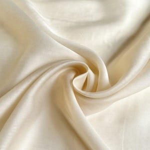 以下が含まれることがあります： 柔らかく流れるような質感のクリーム色の生地のクローズアップ。生地はドレープされ、折りたたまれており、その滑らかな表面を強調する影とハイライトを作り出しています。素材は軽く、豪華に見え、おそらくシルクまたは同様のテキスタイルです。